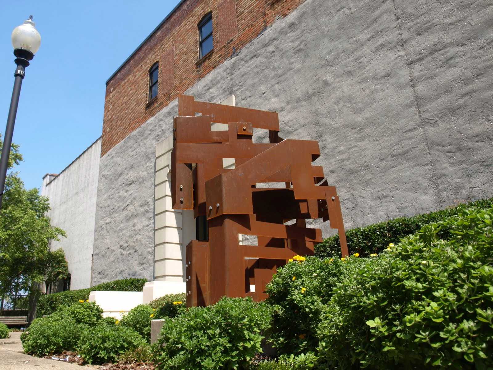 Large sculpture of bent metal bolted together reaching to the sky, integrated into a garden setting